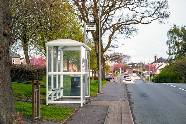 Glass-sided bus shelter beside a quiet suburban street with visible bus stop sign and passing traffic. Spring trees with fresh green leaves and pink blossoms line the pavement.