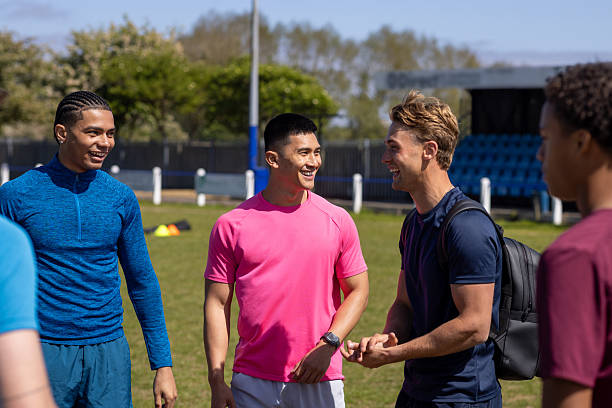 A medium view of a diverse group of young male friends standing together on the football pitch at football training in the North East of England. They are chatting before they start football practice. They are wearing sports clothing and carrying sports equipment.Videos are available for this scenario.