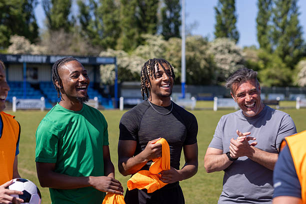 A medium view of a mature football coach talking to a diverse group of young male amateur footballers, they are standing around a table near the football pitch at football training in the North East of England. The coach is giving a pep talk before the team starts football practice. They are wearing sports clothing.Videos are available for this scenario.