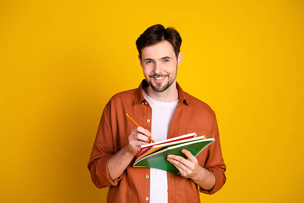 istock Young man with beard holding notebooks and pencil standing against a bright yellow background 2227493575