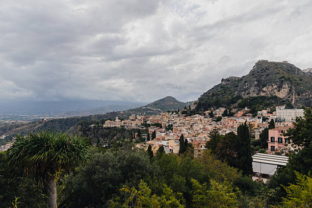 Scenic panoramic view of the hillside town of Taormina in Sicily, Italy, surrounded by lush greenery and rugged mountains under a dramatic cloudy sky.