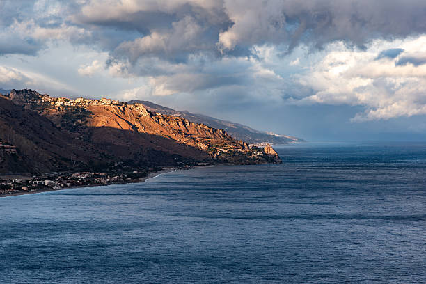 A breathtaking coastal view of sun-drenched cliffs contrasting with dark clouds over the Mediterranean Sea in Sicily, Italy.