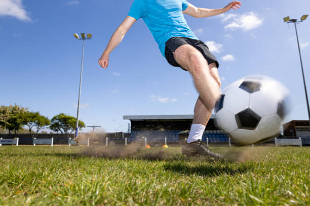 A wide shot of unrecognisable legs in motion as they strike a football on a dusty gravel surface. Small stones and dust scatter through the air, capturing the raw energy and impact of grassroots football. The image focuses on movement and texture, perfect for themes of sport, action, and determination.Videos are available similar to this scenario.
