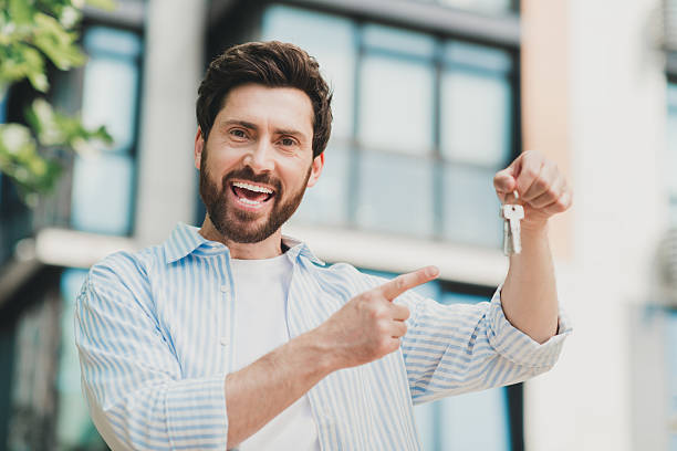 Young bearded man smiling with keys outdoors next to modern building, representing themes of real estate success and happiness