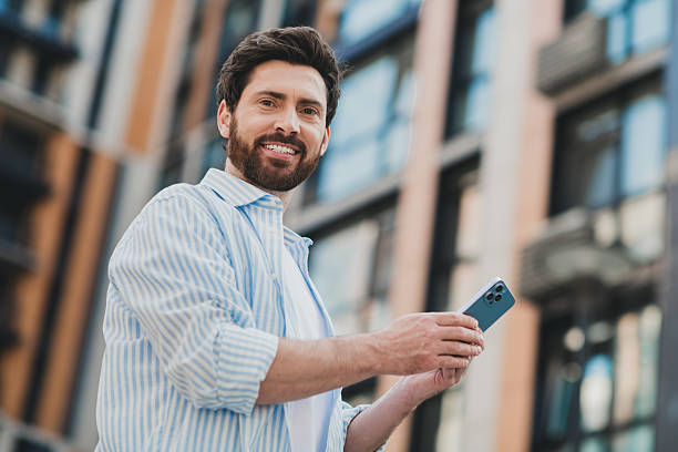 istock Attractive young man with stylish beard using smartphone outdoors near urban buildings under bright sunshine 2227498445
