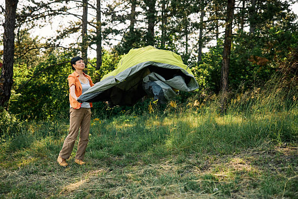 Japanese female camper setting up a green camping tent in a scenic forest, wearing casual outdoor attire under bright daylight, surrounded by tall trees and grass.