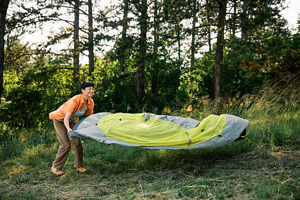 A Japanese woman in casual attire sets up a yellow tent during a peaceful camping trip in a lush forest on a sunny day.