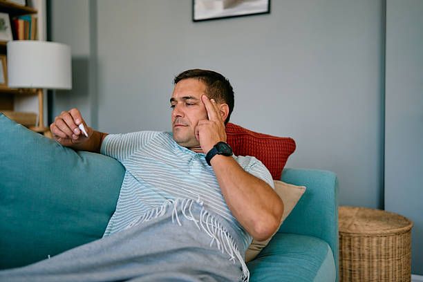 istock Sick man checking his temperature while lying on the sofa at home 2227503122