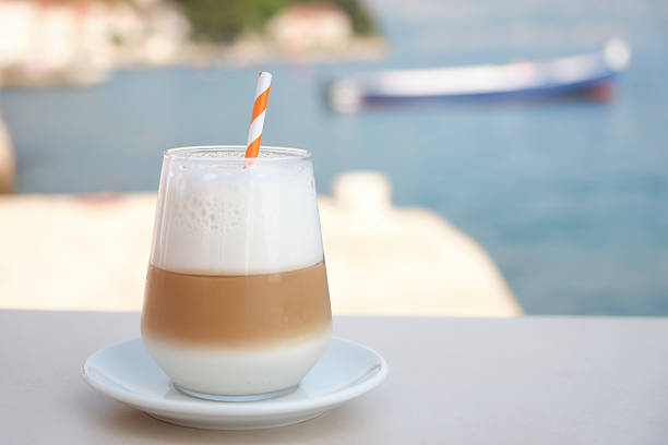 Glass of iced latte with creamy milk foam and an orange striped straw served on a white table overlooking the Adriatic Sea. A small boat is softly blurred in the background, adding to the calm, Mediterranean mood. Ideal for themes like summer, travel, coastal lifestyle, and refreshment. No people, no logos, no property release required.