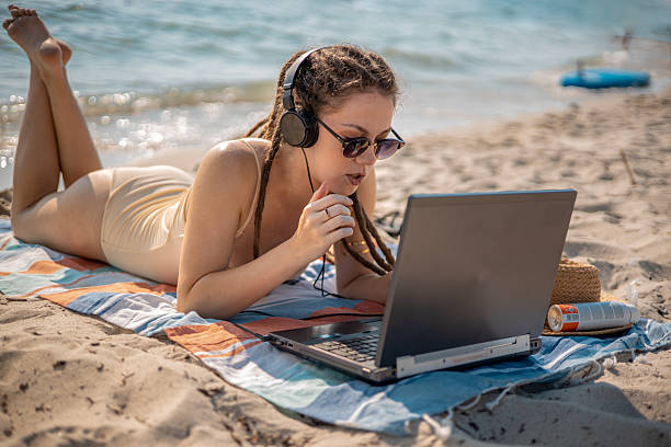 istock Female online language tutor with dreadlocks teaching via laptop and headphones, lying on a beach mat by the sea - remote work lifestyle 2227519953
