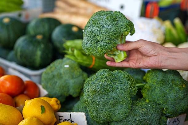 Close-up of a hand holding a fresh broccoli floret over a vegetable display filled with healthy greens and produce.