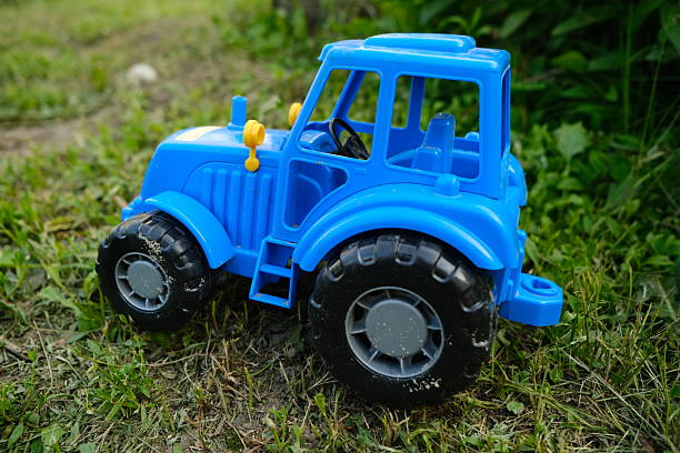 A bright blue toy tractor sits on a patch of green grass surrounded by foliage Sunlight highlights the plastic details showcasing its playful design and vivid colors.
