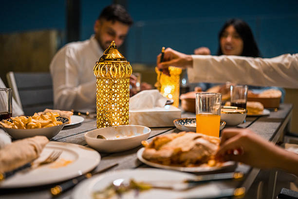 Young Arabic friends gather on a rooftop at night to share a Ramadan dinner. The table is adorned with a glowing lantern, diverse traditional dishes, and drinks, creating a warm and festive atmosphere under the dark sky.