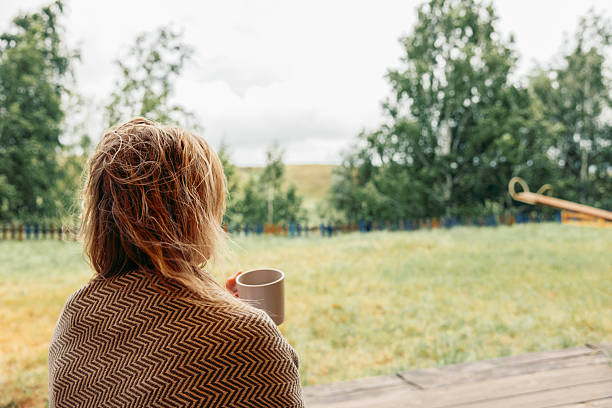 woman drinking warm beverage while looking into distance, rainy outdoor background with natural greens, concept of comfort, calm morning, wellness