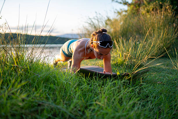 Young athletic woman practicing plank exercise on a mat in a grassy area by the lake at sunset, enjoying a healthy lifestyle