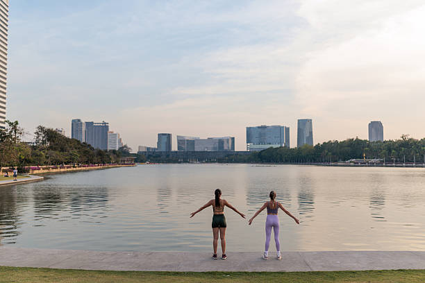 Two female friends exercise side by side in a peaceful park overlooking the Chao Phraya River in Bangkok. Dressed in sportswear and fully focused, they share a calming and empowering moment during their fitness routine.