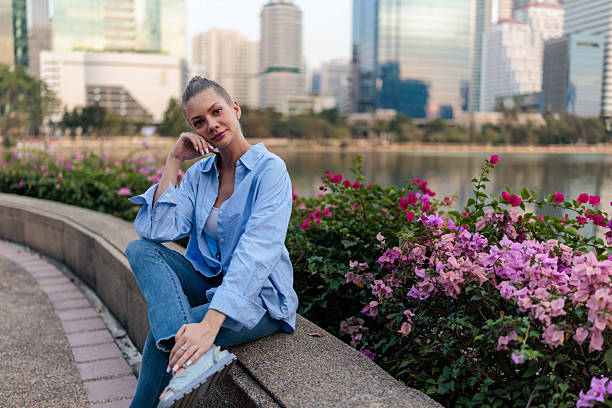 A beautiful, casually dressed woman sits smiling peacefully by the Chao Phraya River in Bangkok’s Benchakitti Park. Surrounded by vibrant pink blossoms, her natural charm and joyful expression create a serene and uplifting moment amid the urban oasis.