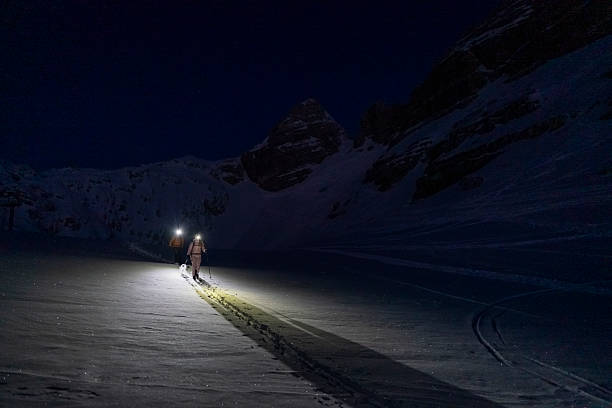 Two skiers with headlamps traverse a serene, snow-covered alpine landscape in the early morning, surrounded by towering peaks under a dark, starry sky.