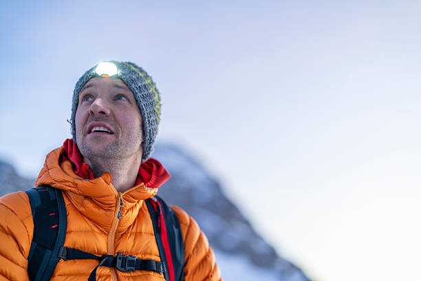 Close-up portrait of mid-aged Caucasian mountaineer in orange down jacket and gray knit beanie, gazing upward with determination against snow-covered mountain backdrop.
