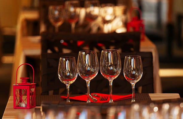 Four empty wine glasses are arranged on a wooden restaurant table alongside a red decorative lantern, with warm ambient lighting and blurred restaurant interior in the background. The scene creates an intimate dining atmosphere perfect for romantic evenings or wine tastings