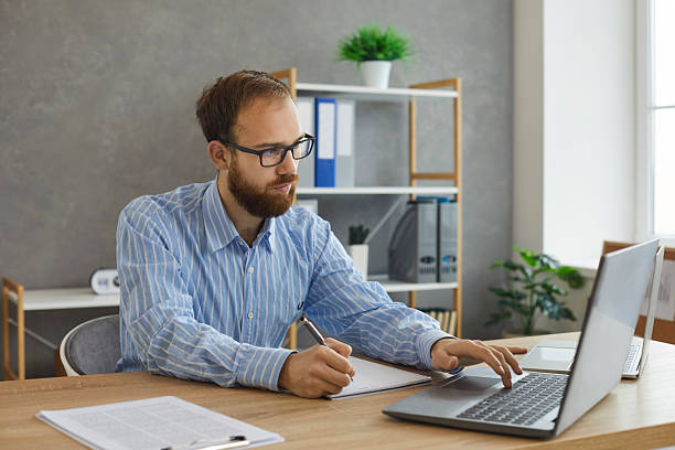 istock Young serious concentrated man working on laptop writing notes on paper sheet 2227572106