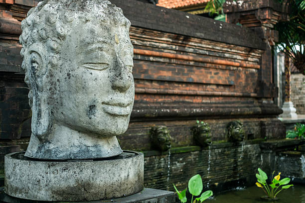 Weathered stone Buddha head statue in a tranquil garden fountain, with traditional Balinese temple walls and greenery in background.