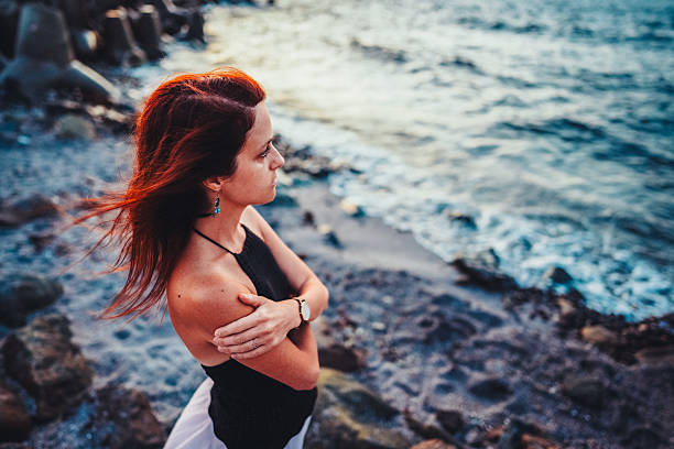 Woman stands by the sea during sunset, gazing thoughtfully at the water with her arms crossed. Her hair is gently blown by the breeze, and she wears a sleeveless black top and flowing skirt. The soft evening light and blurred ocean background create a serene and contemplative mood, capturing a peaceful moment of solitude and reflection by the coast.