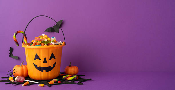 istock Festive Halloween setup featuring a pumpkin-shaped bucket filled with candy on a side view table purple background 2227600356