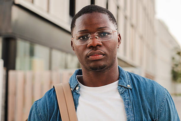 istock Serious African American student looking pensive at the camera, holding a folder and wearing a backpack, standing outdoors in a modern urban setting, expressing confidence at university campus 2227601440