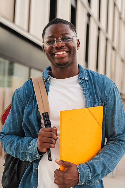istock Vertical. Smiling African American student looking at the camera, holding a folder and wearing a backpack, standing outdoors in a modern urban setting, expressing confidence and positivity. 2227601988