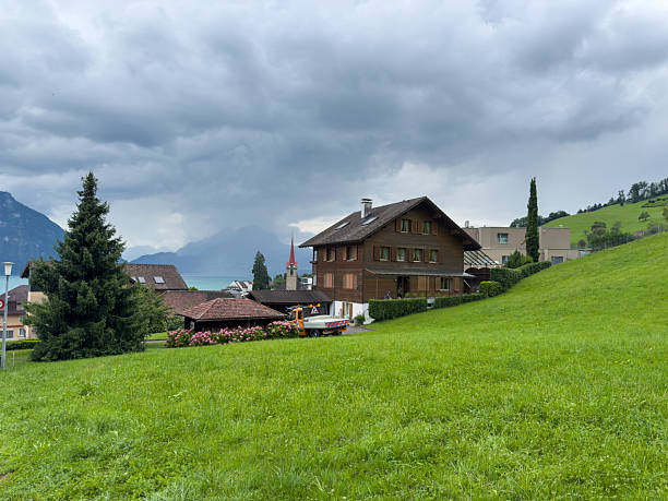 Wooden chalet house with sloped roof on lush green hillside under cloudy sky. Scenic rural village with mountain backdrop and church steeple. Alpine countryside landscape with homes and nature.
