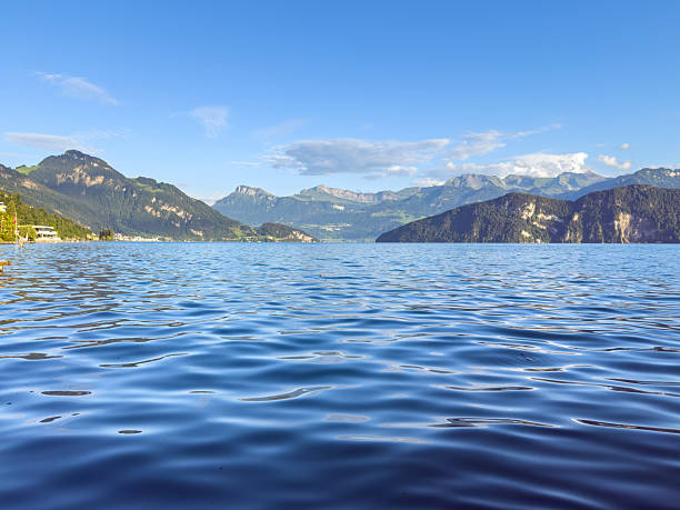 Smooth blue lake water reflecting sunlight with distant green mountains under a clear sky. Serene natural scene of lake and mountain range. Peaceful outdoor landscape with water and hills.