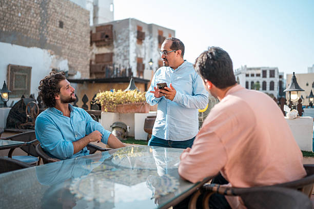 Three men, dressed casually, enjoy dining outdoors under bright sunlight in Al-Balad. The historic architecture and vibrant atmosphere enhance the scene.