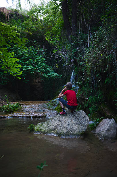 istock Man Seated on a Rock Near a Small Waterfall Surrounded by Lush Green Forest 2227612102