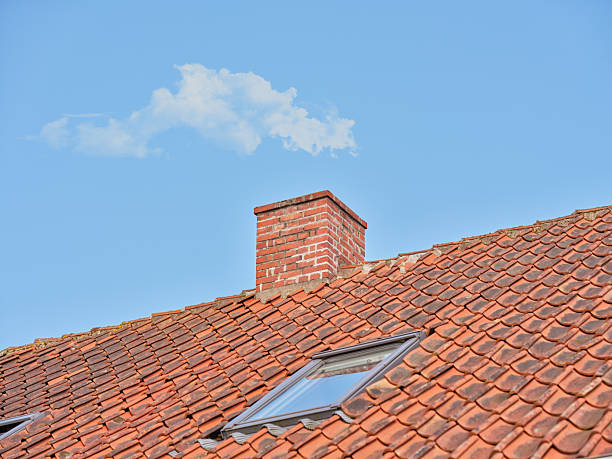 The image showcases a red tiled rooftop featuring a brick chimney and a skylight, with a scattered cloud adorning the pristine blue sky. This charming scene is set in Denmark and evokes serenity and calm.