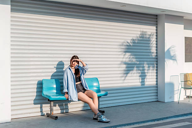 Young woman sitting on blue bench outdoors against roller shutter door, enjoying the sunlight.