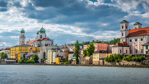 Riverside view of Passau in Bavaria, Germany, featuring colorful Baroque buildings and the green-domed St. Stephen’s Cathedral (Stephansdom). Dramatic summer sky. Passau in lower Bavaria, Germany - Europe