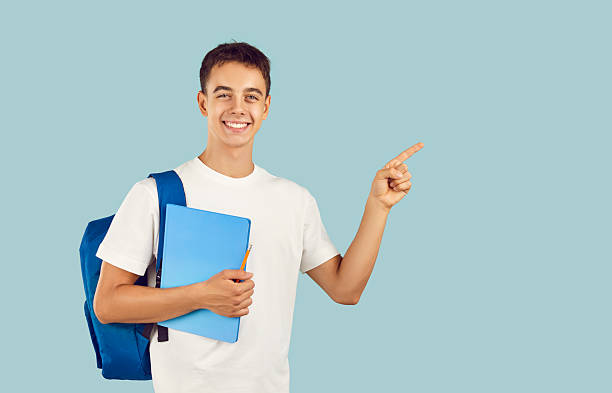 istock Happy boy student holding notebook with backpack pointing finger to the side on blue background. 2227647835