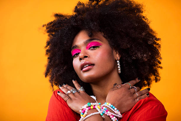 Portrait of at young Black gen z teen girl poses with bold pink eyeshadow and vibrant beaded jewelry, set against a warm yellow background