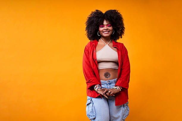 Portrait of a multicultural woman with afro-textured hair, wearing a red shirt, crop top, and ripped jeans, standing in studio with a yellow backdrop, showcasing a stylish and modern aesthetic