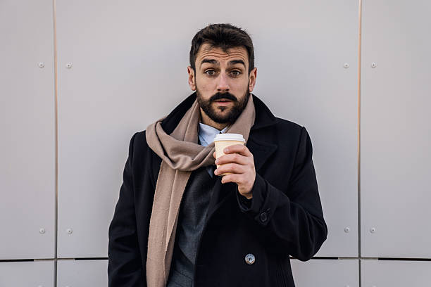 A handsome young entrepreneur dressed warmly holds a hot beverage and gazes with curiosity at the camera while standing outdoors.
