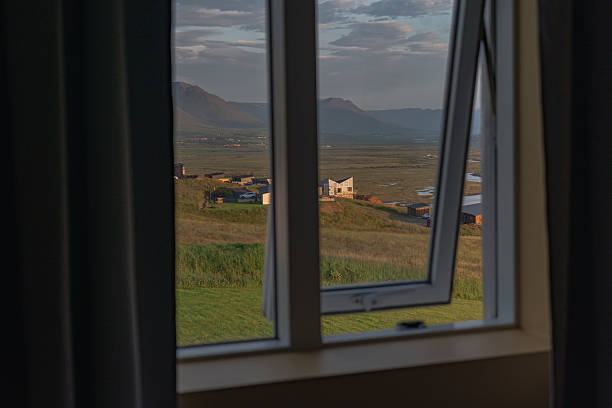 Rolling green fields, a cluster of buildings, and rugged mountains are seen through an open window under a partly cloudy sky with warm sunlight.