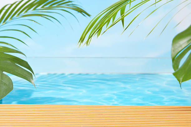 Tropical infinity pool with wooden deck, palm leaves, and bright blue water under clear sky, offering a serene and modern background for vacation or product visuals.