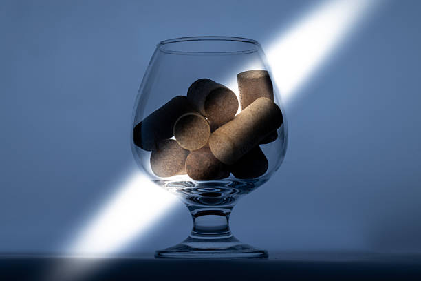 A clear glass, likely a snifter, filled with several natural cork stoppers.A strong beam of light illuminates the glass and corks against a muted
