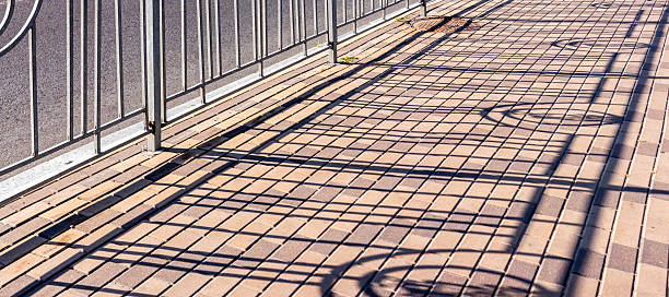 Sunlight casting intricate shadows of a metal fence across a brick paved walkway, forming a captivating geometric pattern enriched with vibrant teal and orange tones