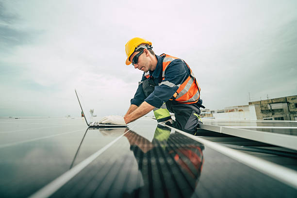 istock Solar technician performing maintenance on photovoltaic panels while using a laptop on a rooftop under a cloudy sky 2227677648