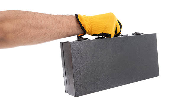 A close-up of a hand wearing a yellow glove, gripping a sturdy black metal case. The background is neutral, emphasizing the action of lifting the case, suggesting movement and purpose.