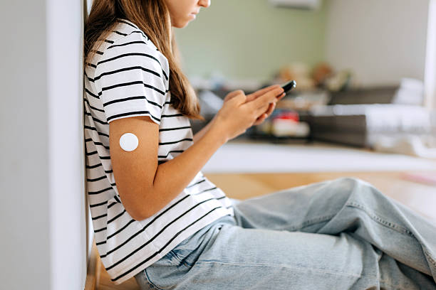 istock Teenage girl with diabetic sensor attached on her arm using smart phone while sitting on floor at home 2227693469