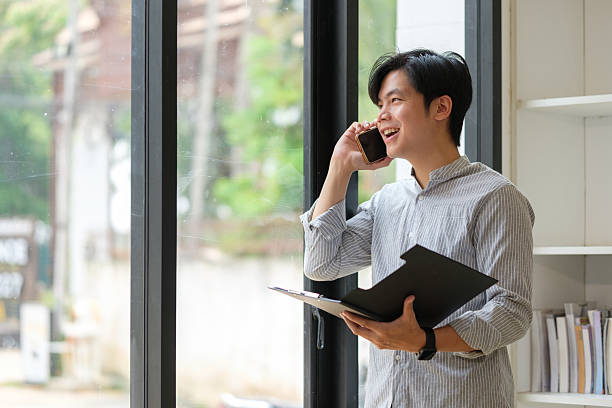 istock Happy office worker making business call while holding clipboard by bright window. 2227695449