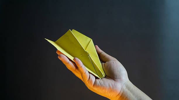 istock Bright yellow paper airplane placed gently on hand, isolated on black background, simple symbolic scene for freedom, childhood, and possibilities 2227696142